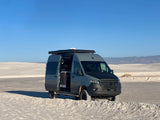 A gray Mercedes Sprinter van, an RV rental, is parked on white sand dunes with its side door open, revealing the interior.