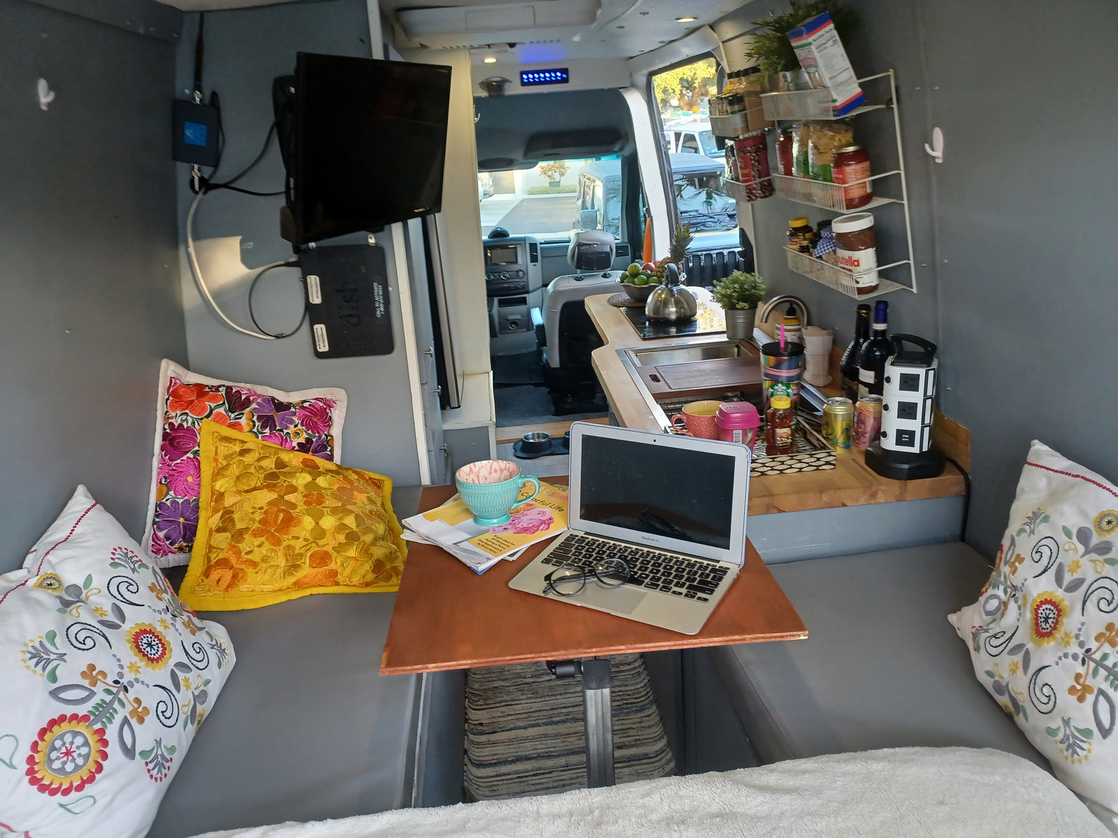 Cozy RV rental interior with a mobile office setup: laptop on table, kitchenette, and comfortable seating with pillows.