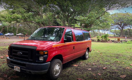 This red Ford van, ideal as an RV rental, is parked on grass in a lush Hawaiian park.