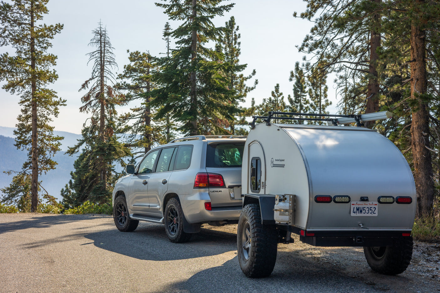 Scenic RV rental: Silver SUV & teardrop trailer parked on a mountain road among tall pines, overlooking mountains.