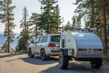 Scenic RV rental: Silver SUV & teardrop trailer parked on a mountain road among tall pines, overlooking mountains.