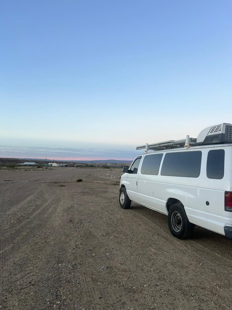 A white RV rental camper van with a roof AC unit, parked on a dirt road in a vast desert landscape at dusk.