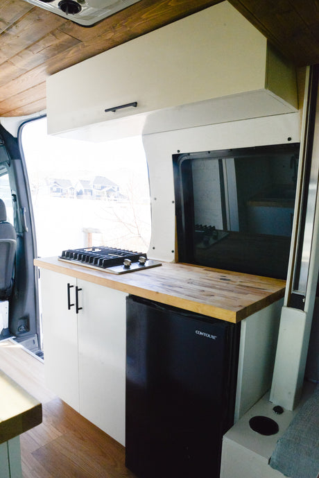 RV rental kitchen: stylish van conversion features a gas stove, wooden counter, mini-fridge, and white overhead cabinets.