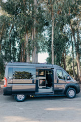 Grey camper van **RV rental** with open side door revealing kitchenette, parked on asphalt among tall trees.