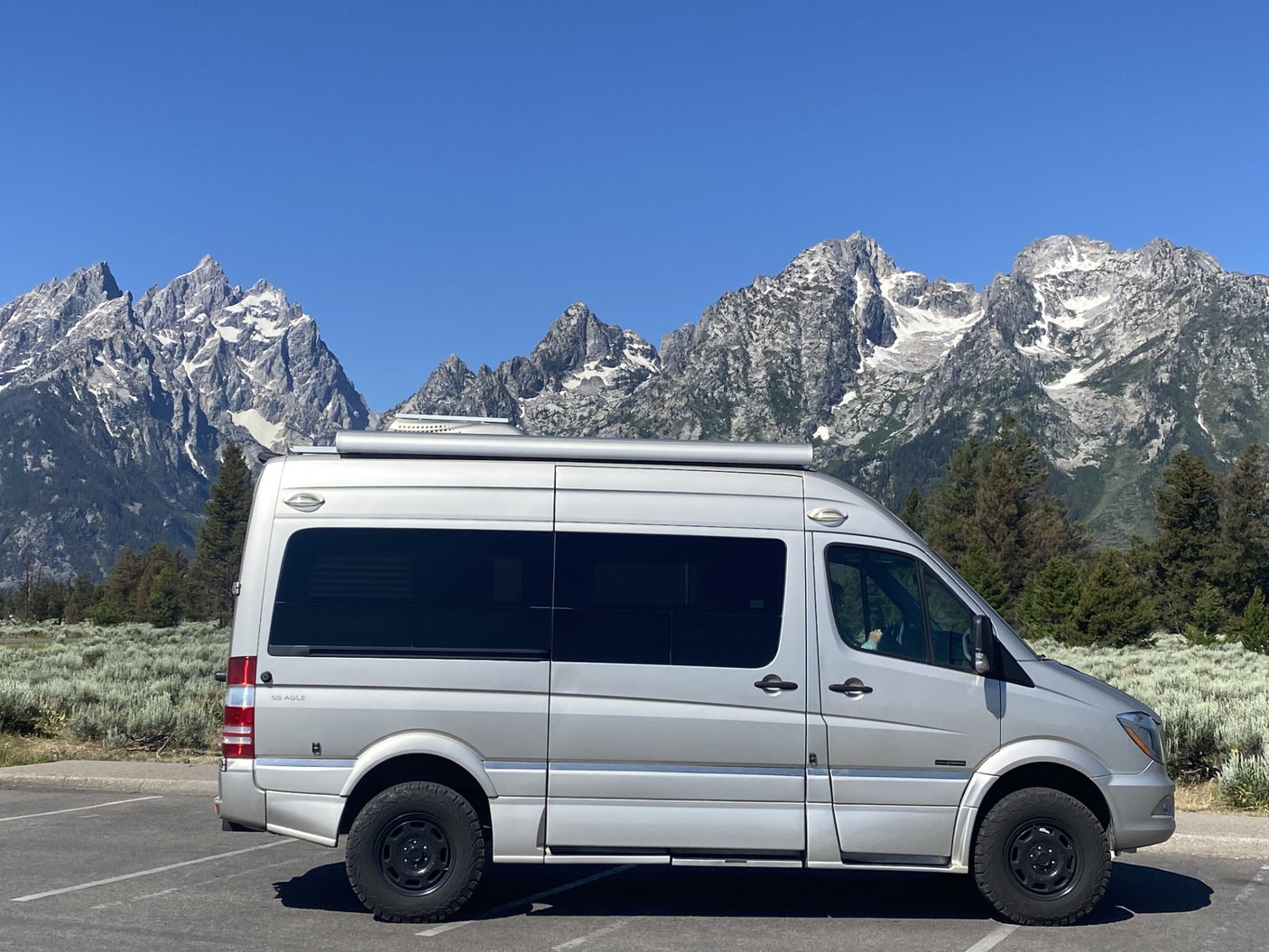A silver Sprinter camper van (RV rental) against the stunning Grand Teton mountains under a clear blue sky.