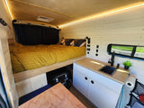 An RV rental's clever interior design shows a comfortable bed, functional kitchenette with sink, wood accents, and bright lighting.
