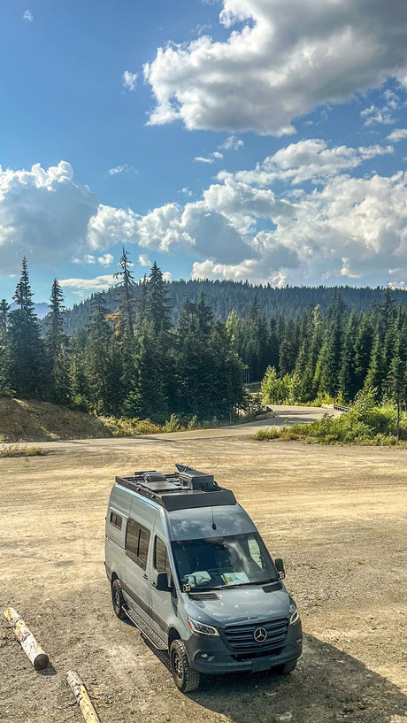 Grey Mercedes Sprinter camper van, perfect for RV rental, parked in a dirt lot with a forest and mountains background.