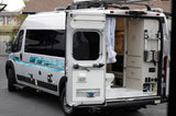 Silver RV rental van with rear doors open, showing the built-in kitchen, toilet, and storage inside, along with exterior utility hookups.