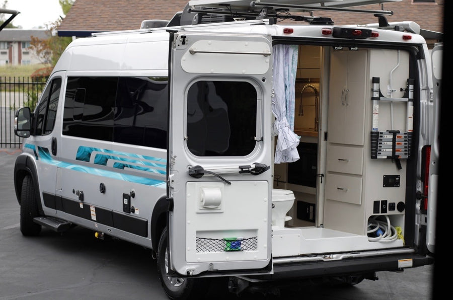 Silver RV rental van with rear doors open, showing the built-in kitchen, toilet, and storage inside, along with exterior utility hookups.