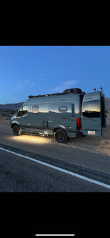 A grey Winnebago Revel 4x4 rv rental parked in the desert at dusk with open doors and side lights on, ready for adventure.