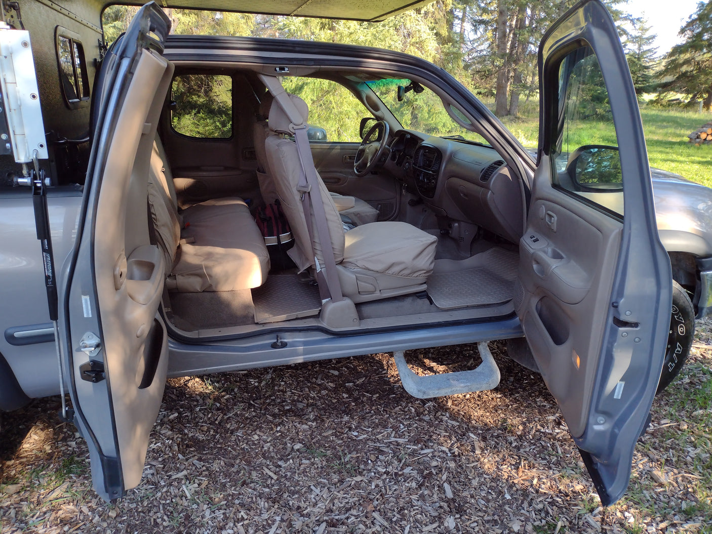 Inside an adventure-ready pickup truck for RV rental, all doors open, showing the spacious tan interior at a forest campsite.