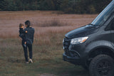 A woman holding a child stands near a dark RV rental van in a grassy field, enjoying family travel and the outdoors.