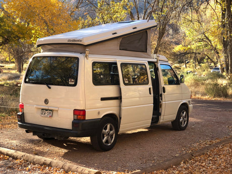 White VW pop-top camper van, a perfect RV rental, parked in a vibrant Colorado autumn setting. Doors open for camping.