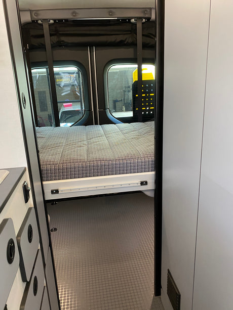 A glimpse inside an RV rental shows a compact sleeping area with a bed, left-side cabinets, and textured floor.