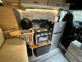 Cozy RV rental kitchen: compact two-burner stove with oven, sink, and mini-fridge. Perfect for your next road trip adventure.