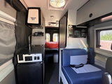 Step inside this RV rental: a compact interior features a kitchenette, controls, blue seating, and a cozy bed in the rear.