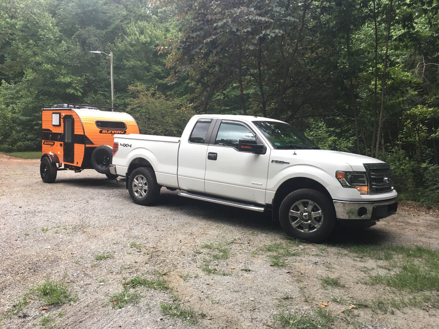 White Ford F-150 pickup truck towing an orange Sunray RV rental on a gravel road, surrounded by trees.