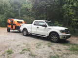 White Ford F-150 pickup truck towing an orange Sunray RV rental on a gravel road, surrounded by trees.
