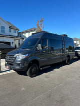 Dark grey Sprinter **RV rental** parked on a street. This rugged **camper van** has off-road tires, a roof rack, and a side ladder.