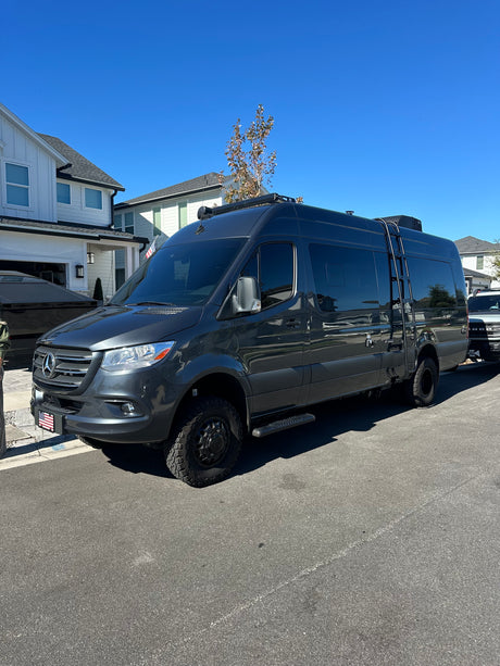 Dark grey Sprinter **RV rental** parked on a street. This rugged **camper van** has off-road tires, a roof rack, and a side ladder.