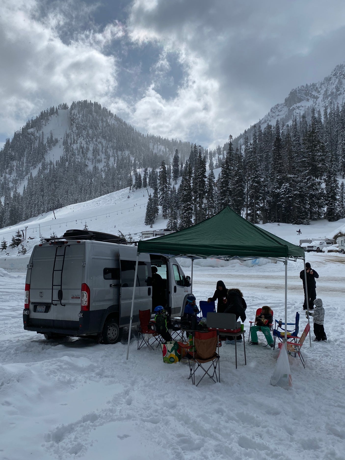 A grey RV rental van and green canopy are set up in a snowy mountain ski resort parking lot. A family relaxes nearby, enjoying the winter scene.
