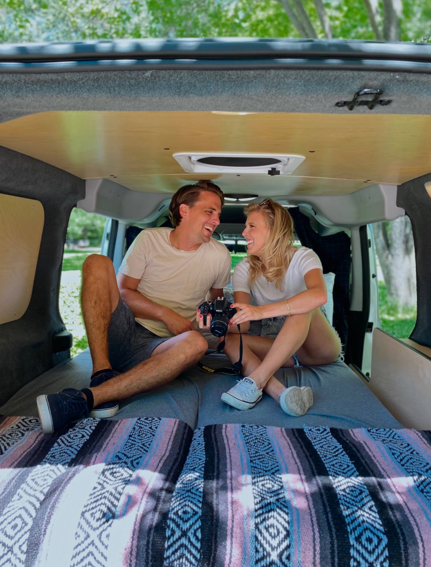 An RV rental's interior: a smiling couple sits comfortably in their camper van, camera in hand, surrounded by wood and blankets. — photo 2