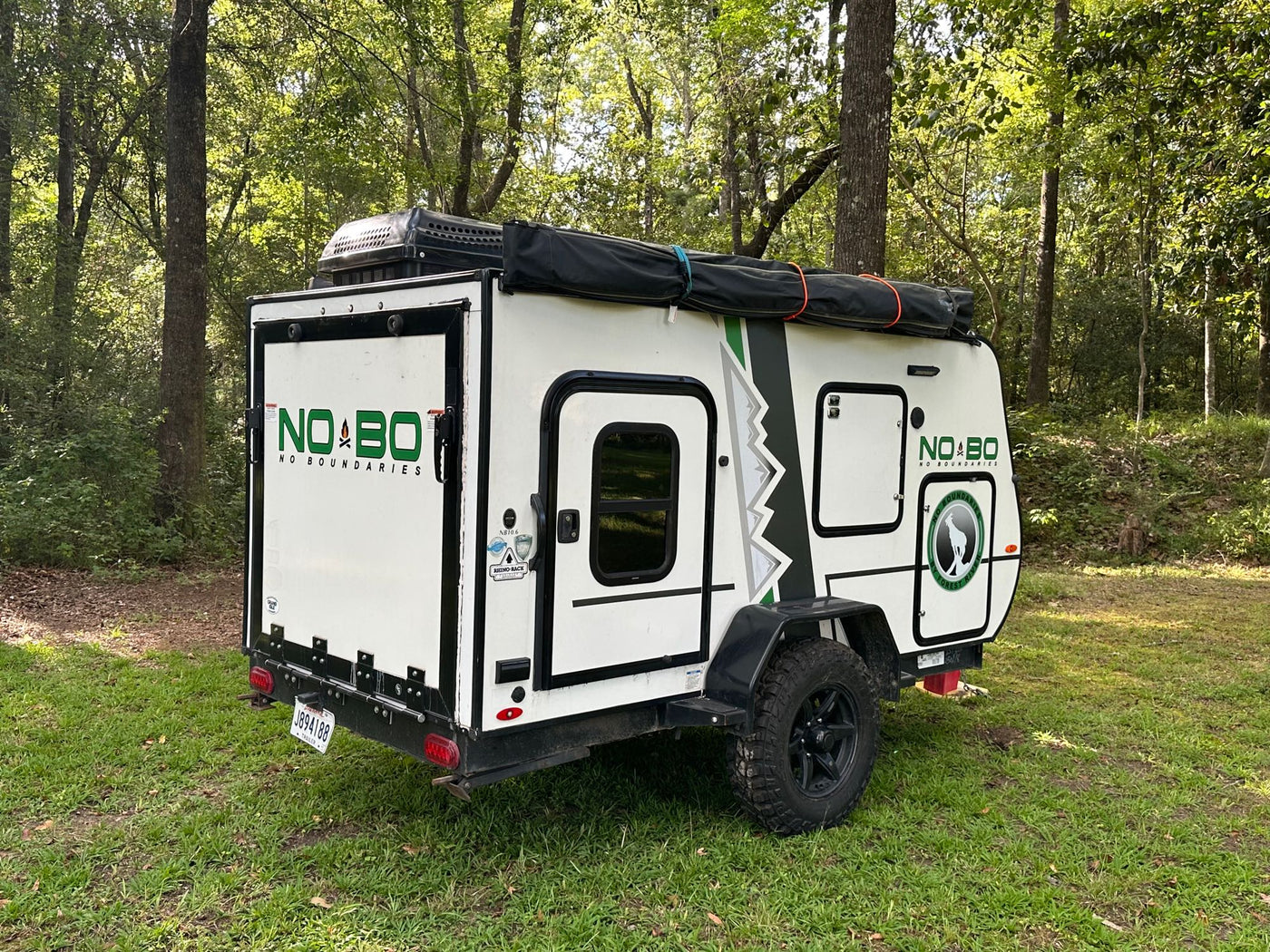 A white NO BO RV rental, a compact camping trailer, is parked on green grass surrounded by trees, ready for adventure. — photo 6