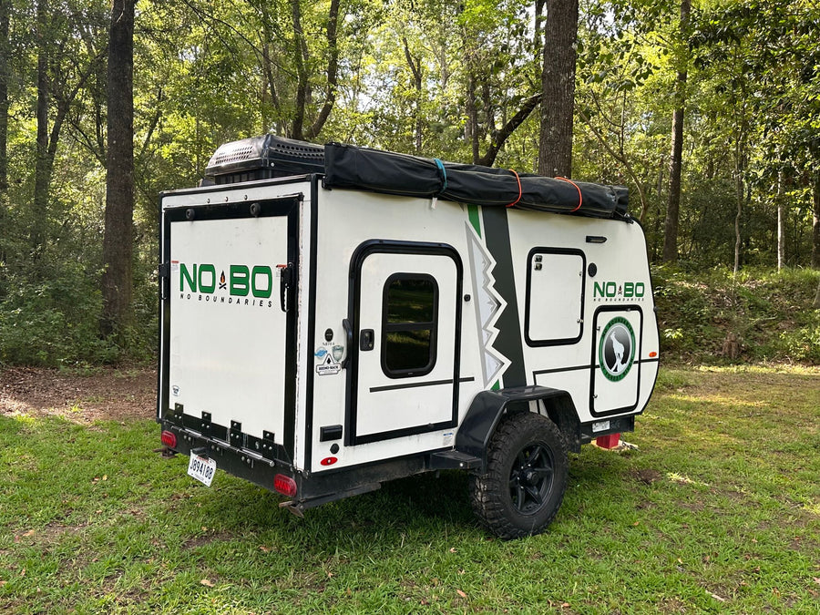 A white NO BO RV rental, a compact camping trailer, is parked on green grass surrounded by trees, ready for adventure.