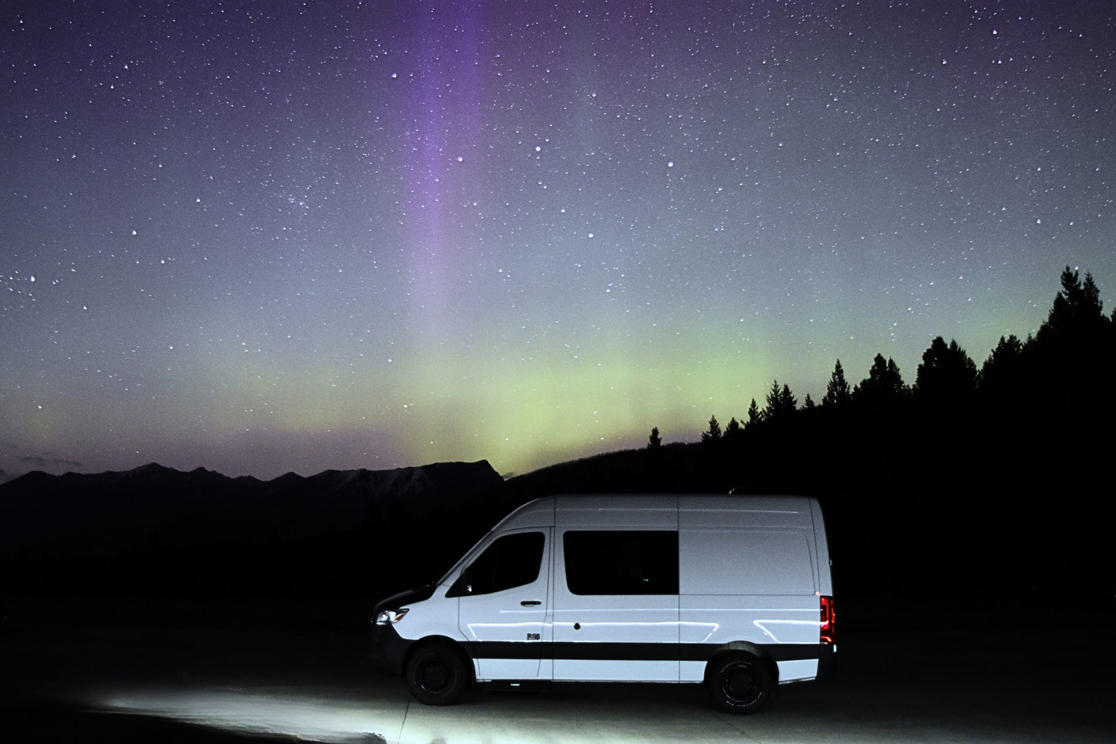 A white RV rental van illuminated under vibrant purple/green aurora, starry sky, silhouetted mountains & trees. Epic adventure awaits.