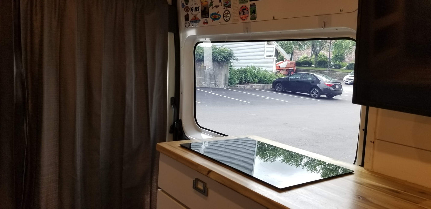 RV rental interior: cozy curtains, counter with cooktop reflecting trees, and a window view of a parking lot.