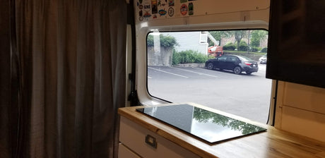 RV rental interior: cozy curtains, counter with cooktop reflecting trees, and a window view of a parking lot.