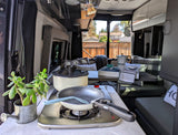 Cozy RV rental interior with a compact kitchen and dining area. Features include a stove, pan, pot, moka pot, and potted plant.