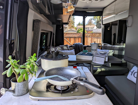 Cozy RV rental interior with a compact kitchen and dining area. Features include a stove, pan, pot, moka pot, and potted plant.