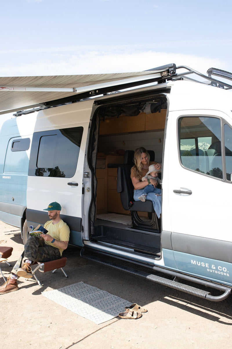 parked on a dirt track amidst trees for outdoor adventures.|Custom blue & white RV rental van with extended awning & rear bike rack — photo 2