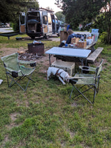 RV rental campsite with an open van, person reading at a picnic table, bulldog, and fire pit. Enjoying the outdoors.