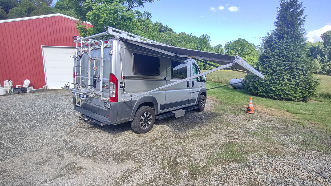 Grey camper van RV rental with extended awning, parked on gravel beside a red barn, ready for outdoor trips.
