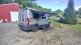 Grey camper van RV rental with extended awning, parked on gravel beside a red barn, ready for outdoor trips.