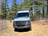 Outdoorsy RV rental: a grey Mercedes-Benz Sprinter camper van parked on a dirt road under tall pine trees.