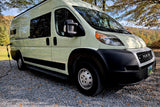 Light green Ram ProMaster camper van (RV rental) parked on gravel, with a power cable, autumn trees, and hills in the background.