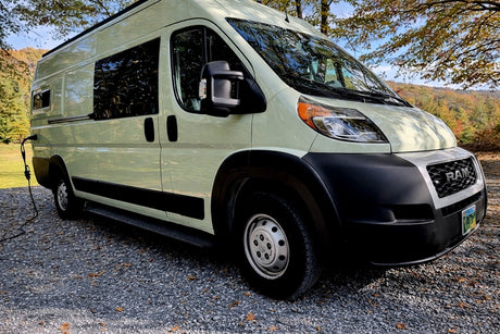 Light green Ram ProMaster camper van (RV rental) parked on gravel, with a power cable, autumn trees, and hills in the background.