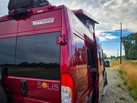 Rear view of a red Winnebago Solis pop-top camper van by a rural road. Perfect RV rental for road trips!