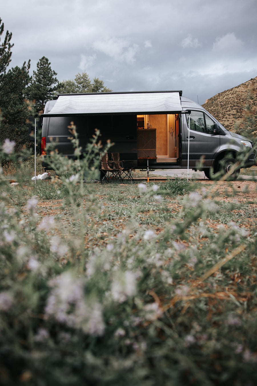 Cozy dark grey RV rental with an extended awning and open, lit interior. Camping chairs invite outdoor relaxation in nature.