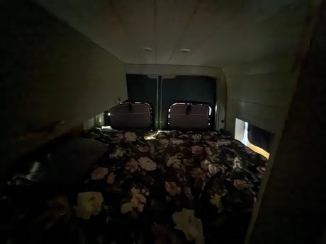 Cozy RV rental sleeping area at night. Dimly lit camper van interior with floral bedding & back window outlines visible.