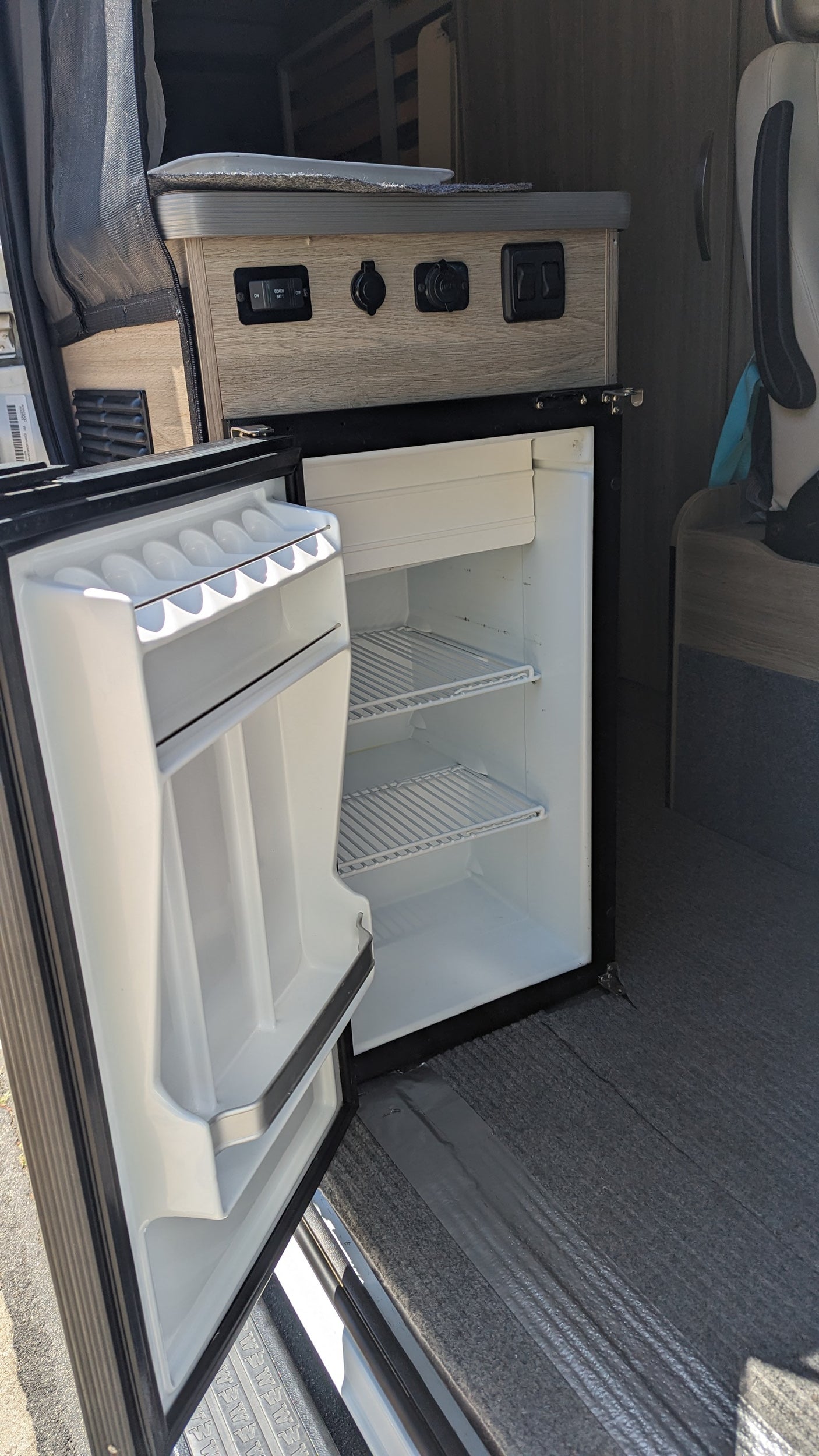 Compact refrigerator with shelves and countertop controls inside an rv rental camper van. Essential for mobile living.