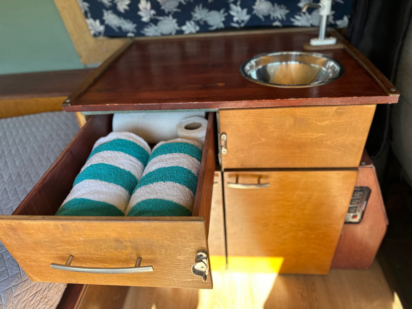 RV rental kitchenette: Wooden counter with sink. Open drawer reveals striped towels & paper towels. Compact for travel needs.