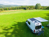 White camper van RV rental with solar panels & bikes, parked in a scenic green field for outdoor adventures.