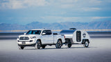 White pickup truck and shiny teardrop RV rental on salt flats with mountains under a blue sky.