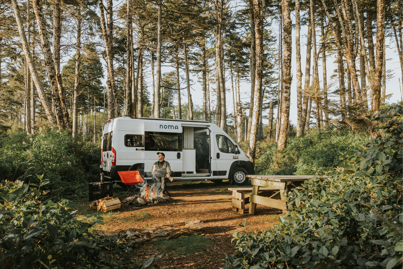 Experience van life with an RV rental! A man sits by a roaring campfire beside his white camper van, surrounded by tall trees. — photo 3
