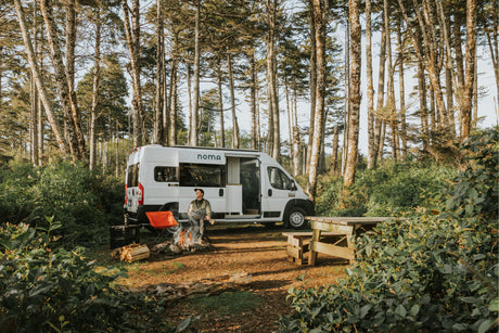 Experience van life with an RV rental! A man sits by a roaring campfire beside his white camper van, surrounded by tall trees.