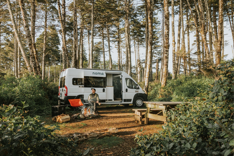 Experience van life with an RV rental! A man sits by a roaring campfire beside his white camper van, surrounded by tall trees. — photo 3